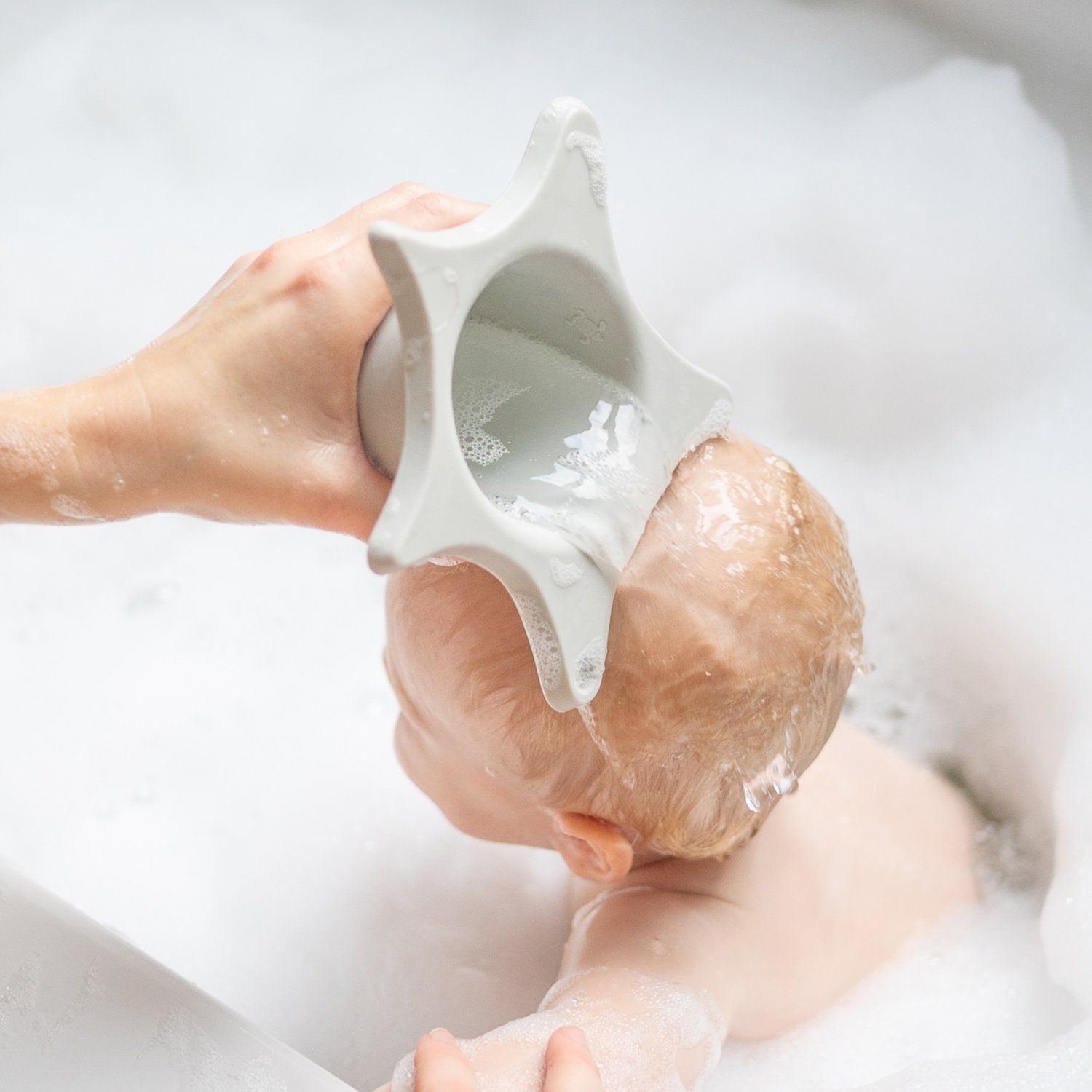 Person hält hellgraue Duschhilfe über dem Kopf eines Babys in einem Waschbecken mit Seifenschaum. Wasser fließt.