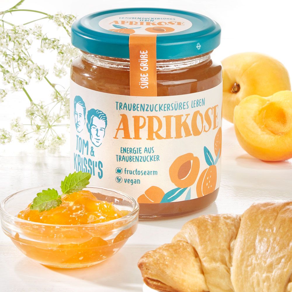 Glas mit Aprikosenmarmelade, daneben eine Schale mit Marmelade und ein Croissant. Aufschrift "Aprikose", "Tom & Krissi's".