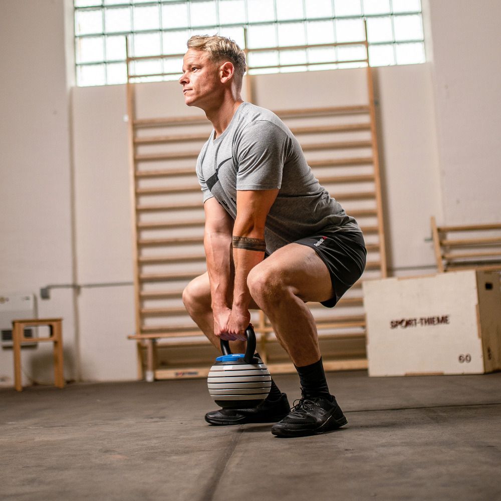 Kettlebell Vario, schwarz-weiß, in Gebrauch. Athlet in tiefer Hocke, Griff gehalten. Hintergrund: Fitnessraum.