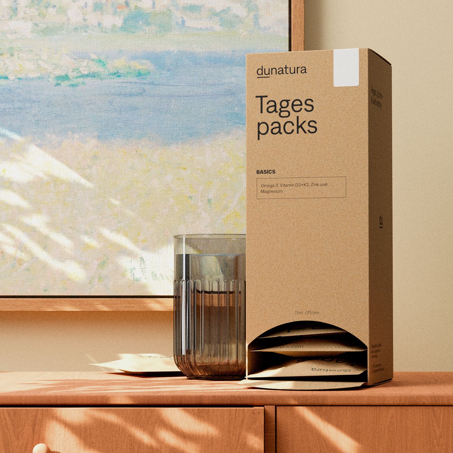Kartonverpackung "Tagespacks" neben Glas Wasser. Aufdrucke: "dunatura", "Basics", Inhaltsstoffe. Holzoberfläche, Kunstwerk im Hintergrund.