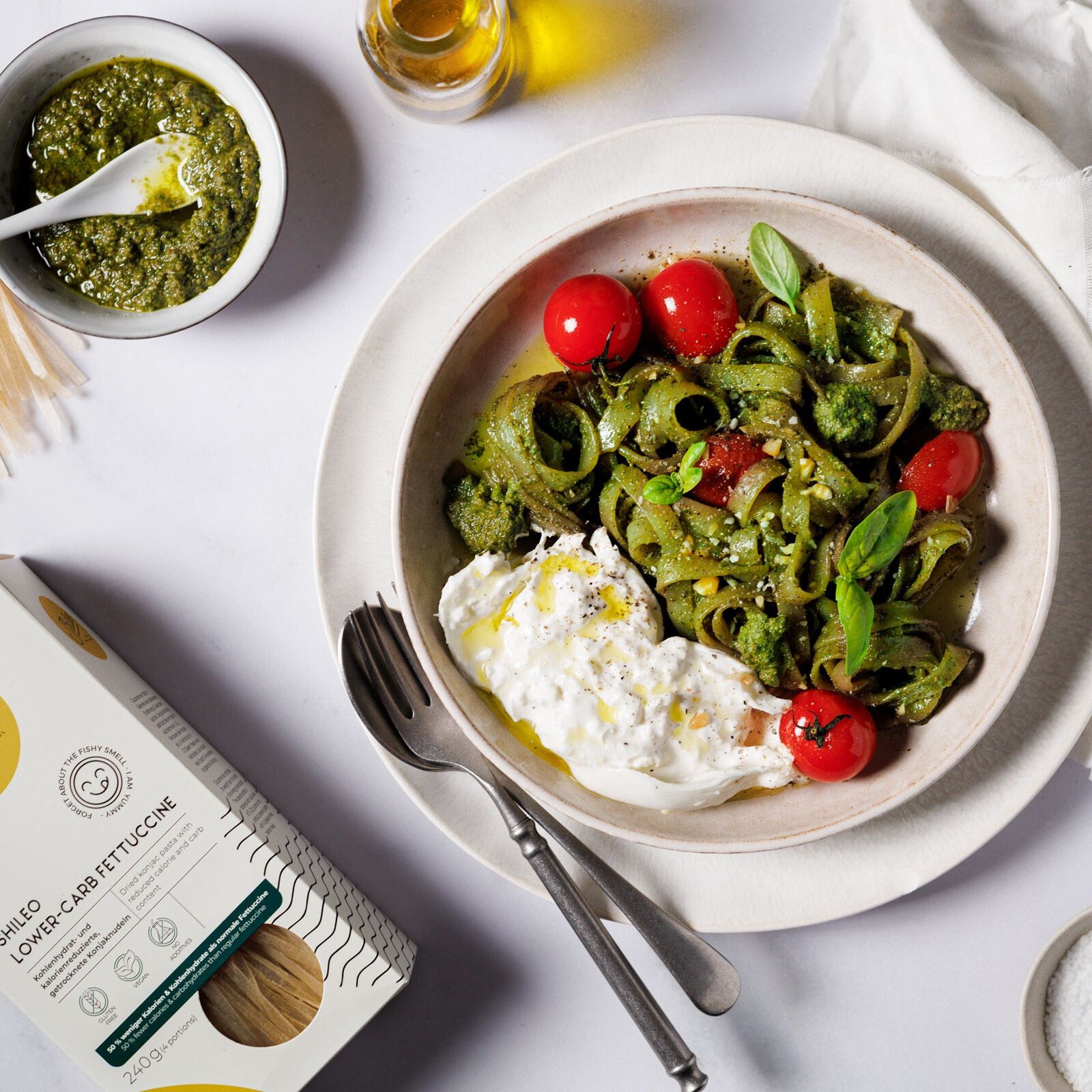 Teller mit grün gefärbten Nudeln, Tomaten, Burrata und Verpackung SHILEO Lower-Carb Fettuccine. Aufdrucke: Logo, Produktname, 240g.