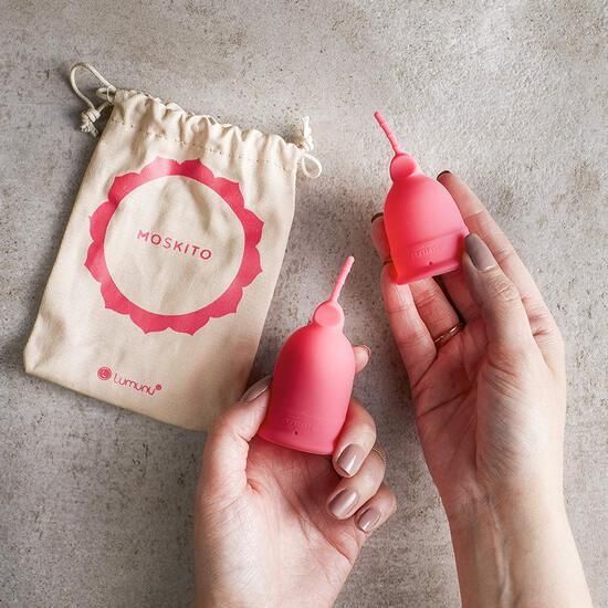 Zwei pinkfarbene Menstruationstassen. Eine Hand hält eine Tasse.