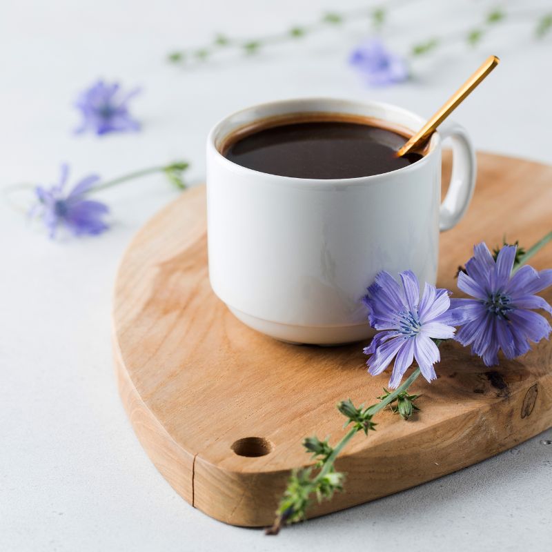 Weiße Tasse Kaffee auf Holzbrett. Goldener Löffel. Blaue Blüten und Zweige umgeben die Tasse.
