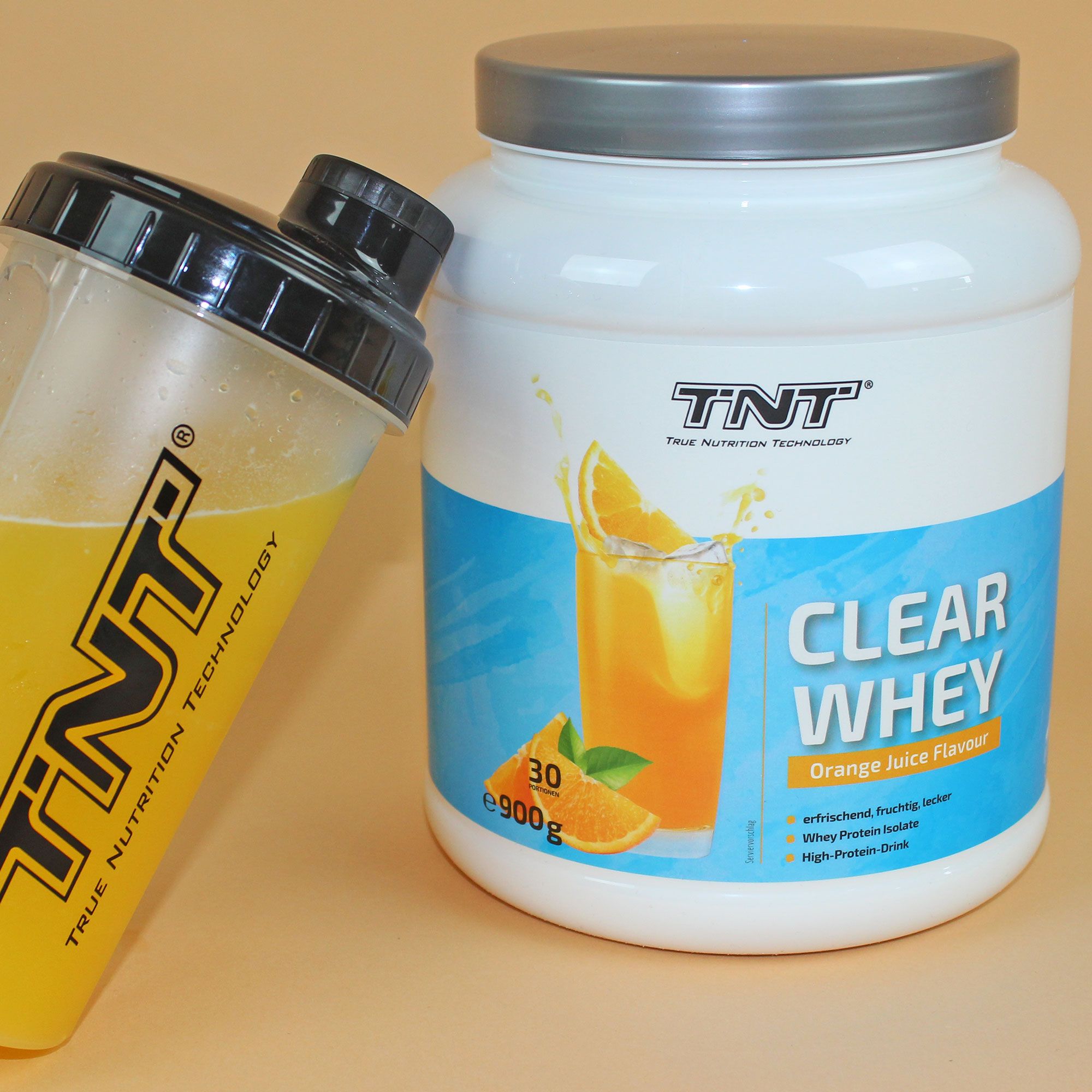 Whey-Protein-Behälter und Shaker. Behälter: weiß, blaues Etikett, Aufdruck CLEAR WHEY. Shaker: schwarz, gelbe Flüssigkeit. TNT-Logo.