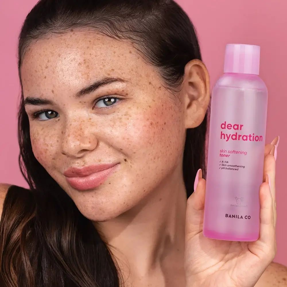 Frau hält rosa Flasche mit "dear hydration"-Aufdruck. Text: "skin softening toner", "A-HA", "pH balanced". Marke: BANILA CO.