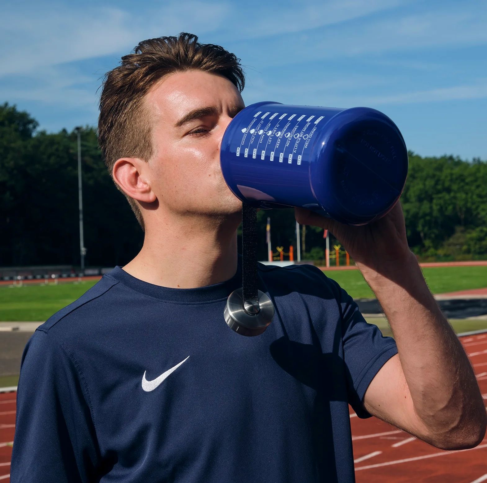 Mann trinkt aus blauer Wasserflasche. Flasche mit Zeitmarkierungen. Metallverschluss. Mann trägt blaues Shirt mit Nike-Logo.