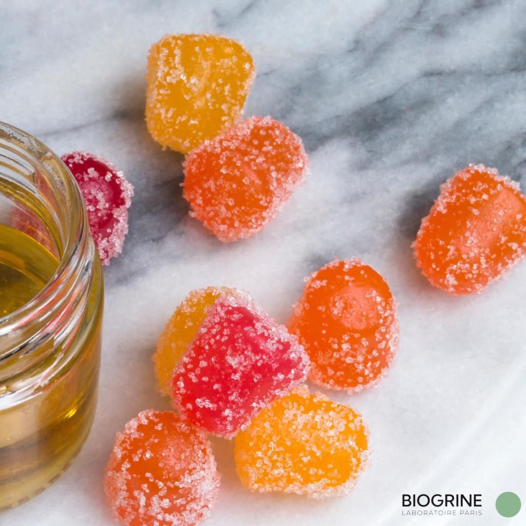 CBD Gummies auf einer Marmoroberfläche. Farben: rot, orange, gelb. Ein Glasbehälter mit Flüssigkeit im Hintergrund.