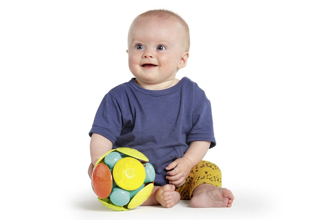 Baby sitzt und hält einen bunten Ball. Der Ball hat gelbe, blaue, orange und grüne Elemente. Das Baby trägt ein blaues Shirt.