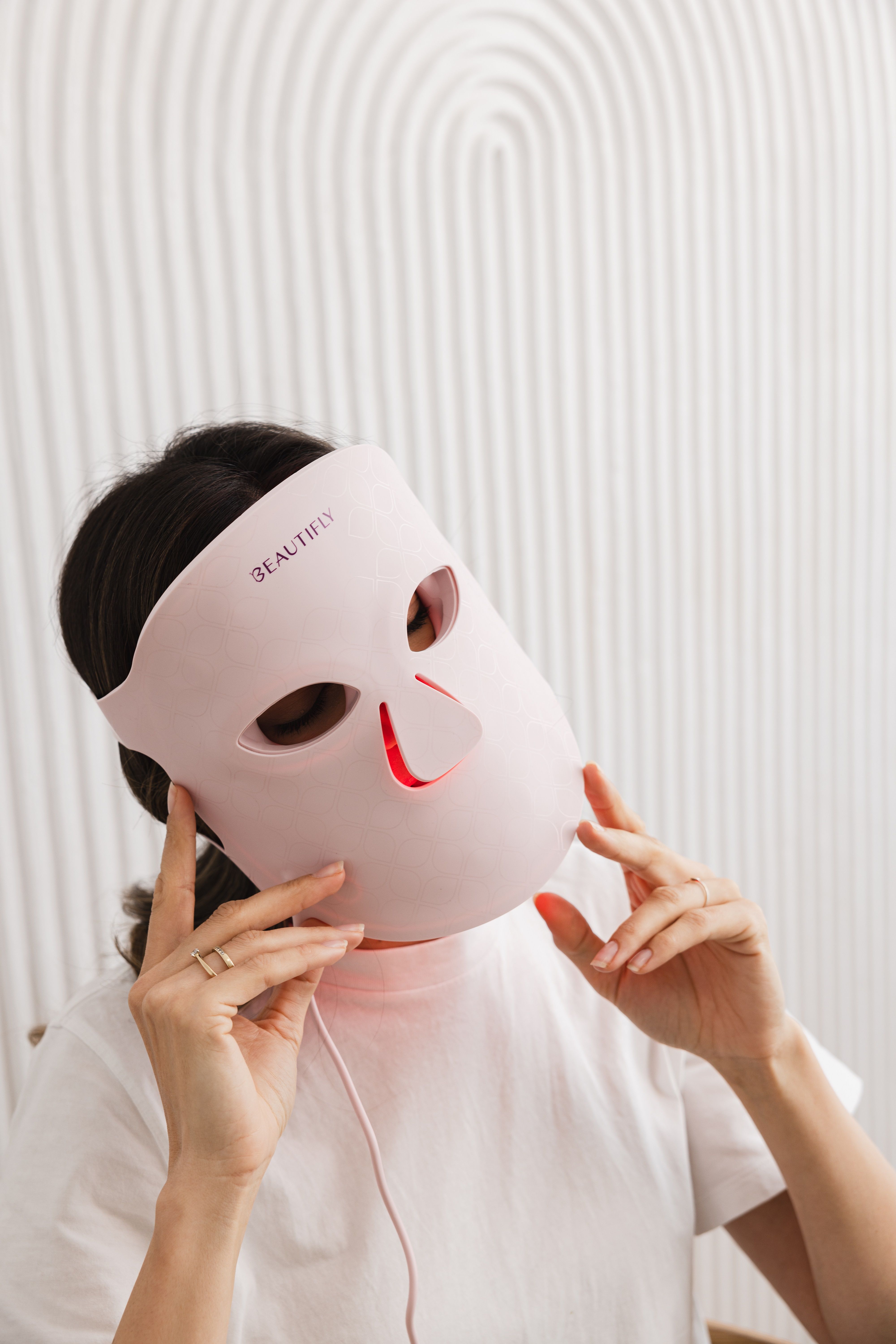 Person trägt eine rosa LED-Gesichtsmaske. Die Maske leuchtet rot. Schriftzug BEAUTIFLY sichtbar.