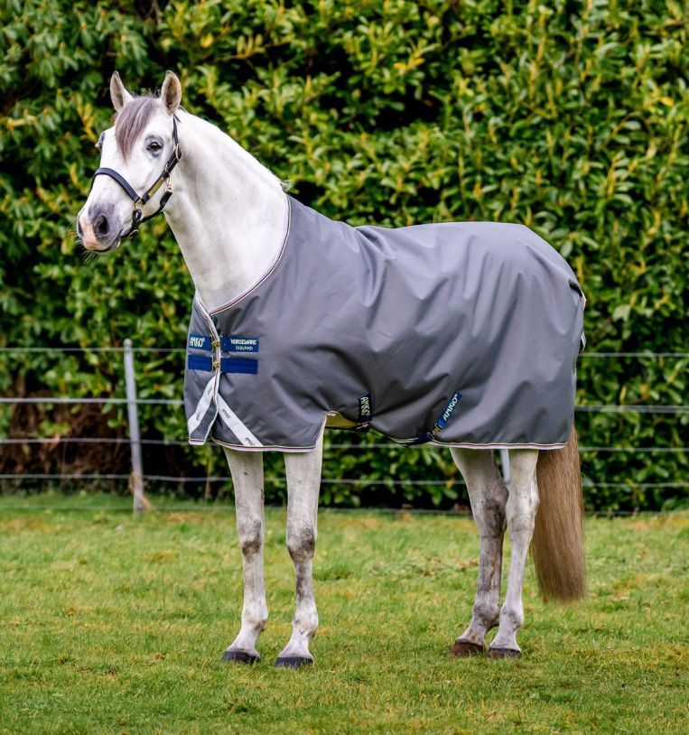 Graue Pferdedecke, getragen von einem weißen Pferd auf einer Wiese. Blaue und weiße Details. Marke: Horseware Amigo.