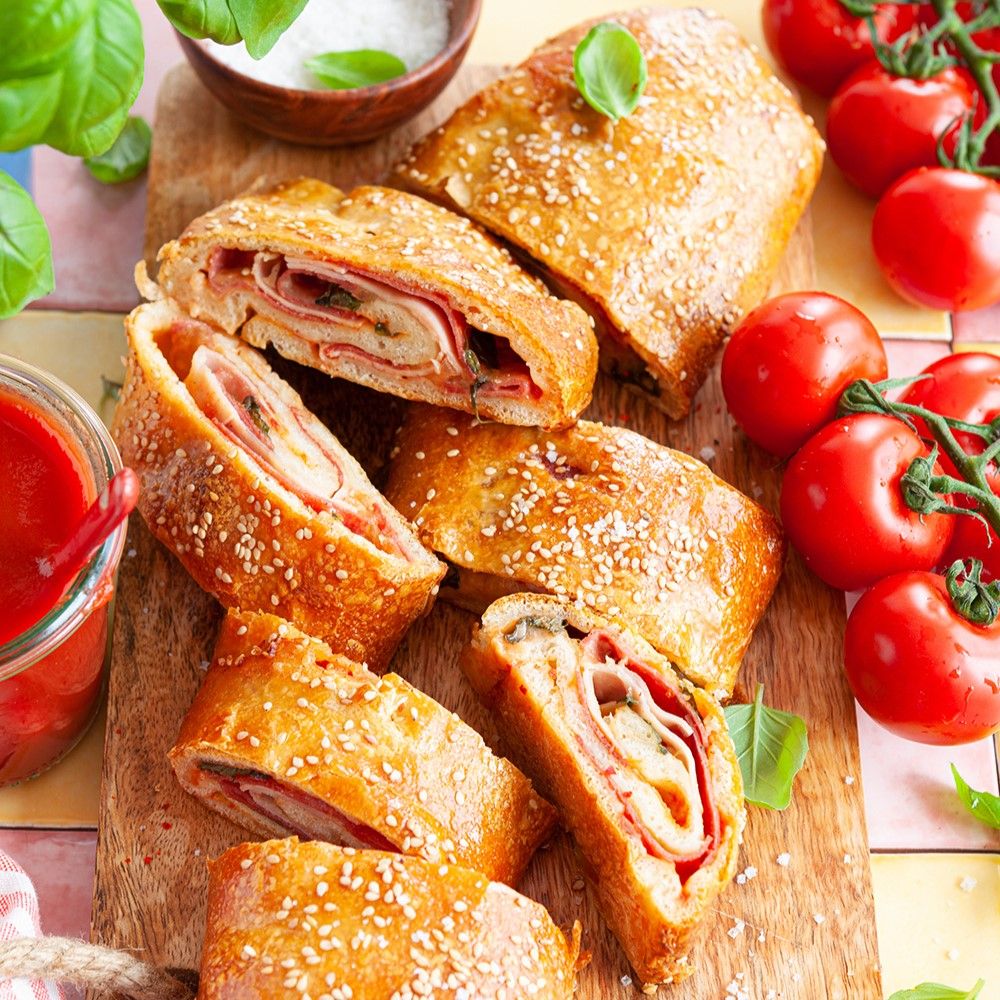 Gebäckstücke mit Füllung auf Holzbrett. Tomaten, Basilikum und Sauce daneben. Mit Sesam bestreut.