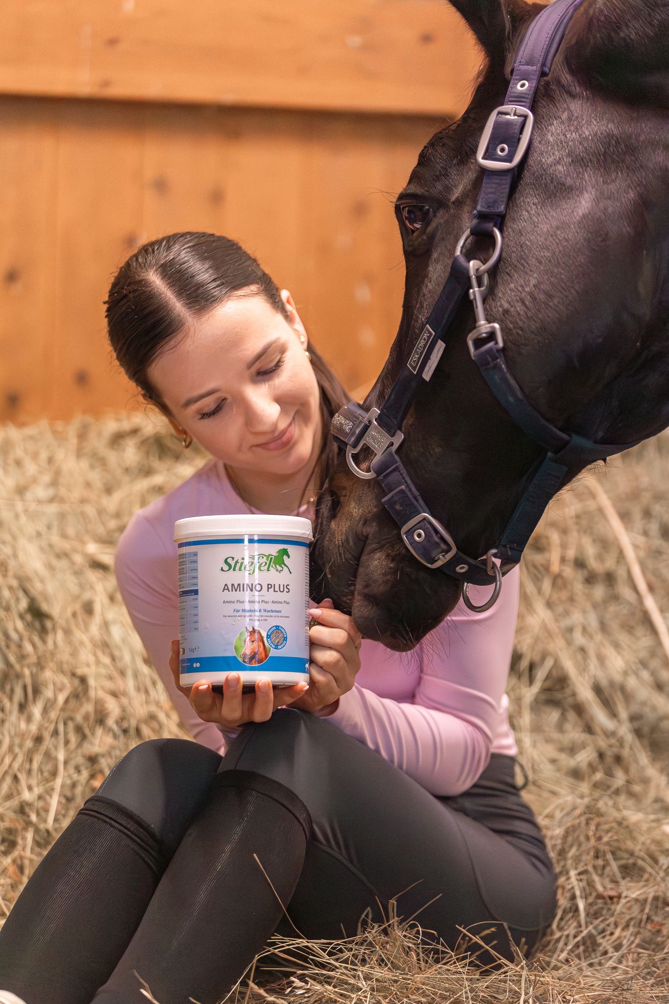 Frau hält einen weißen Behälter mit der Aufschrift Stiefel Amino Plus. Ein Pferd berührt den Behälter mit der Schnauze.