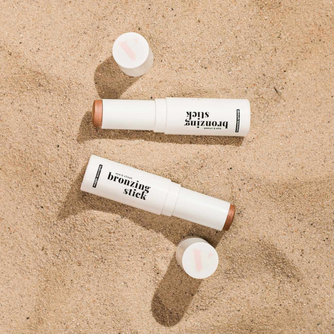 Zwei weiße Bronzing Sticks mit brauner Spitze und schwarzer Schrift auf Sand. Deckel liegen daneben.