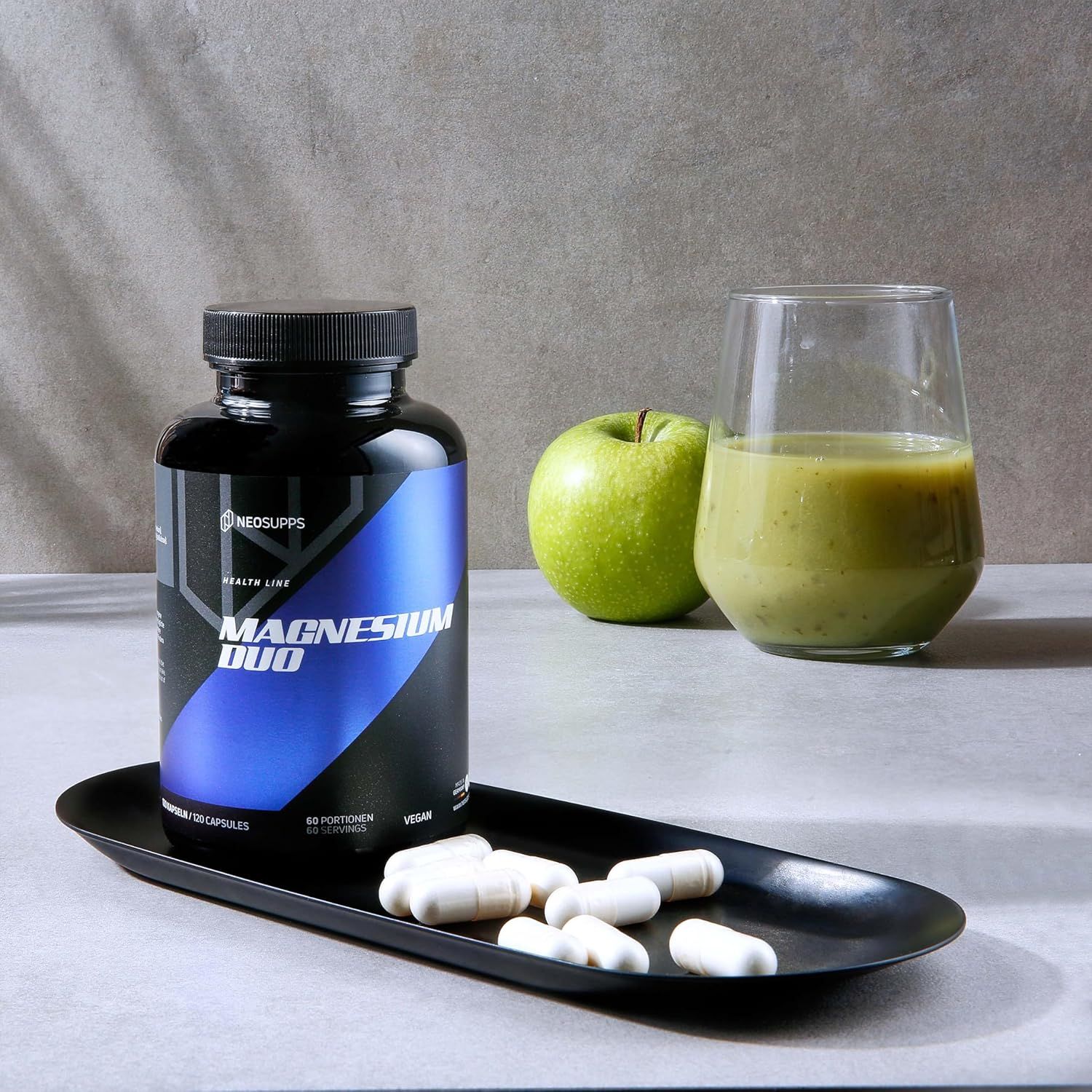 Eine schwarze Tablett mit einer Flasche Neosupps Magnesium Duo, Tabletten, einem grünen Apfel und einem Glas Smoothie.