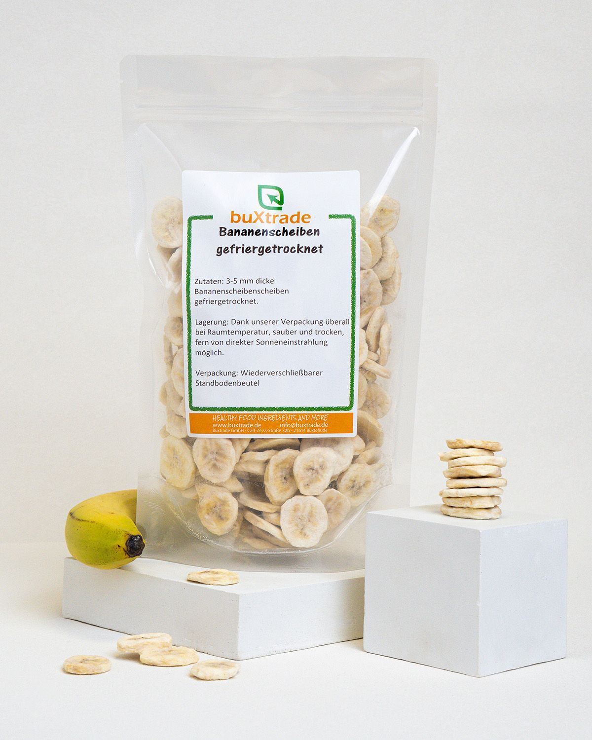 Verpackung von buXtrade Bananenscheiben, daneben eine Banane und ein Stapel Scheiben auf einem Würfel.