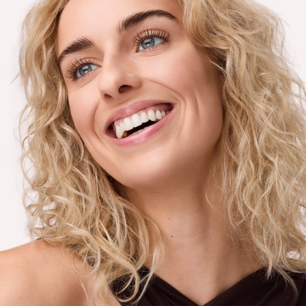 Porträt einer lächelnden Frau mit blonden Locken und blauen Augen. Wimpern scheinen getuscht.