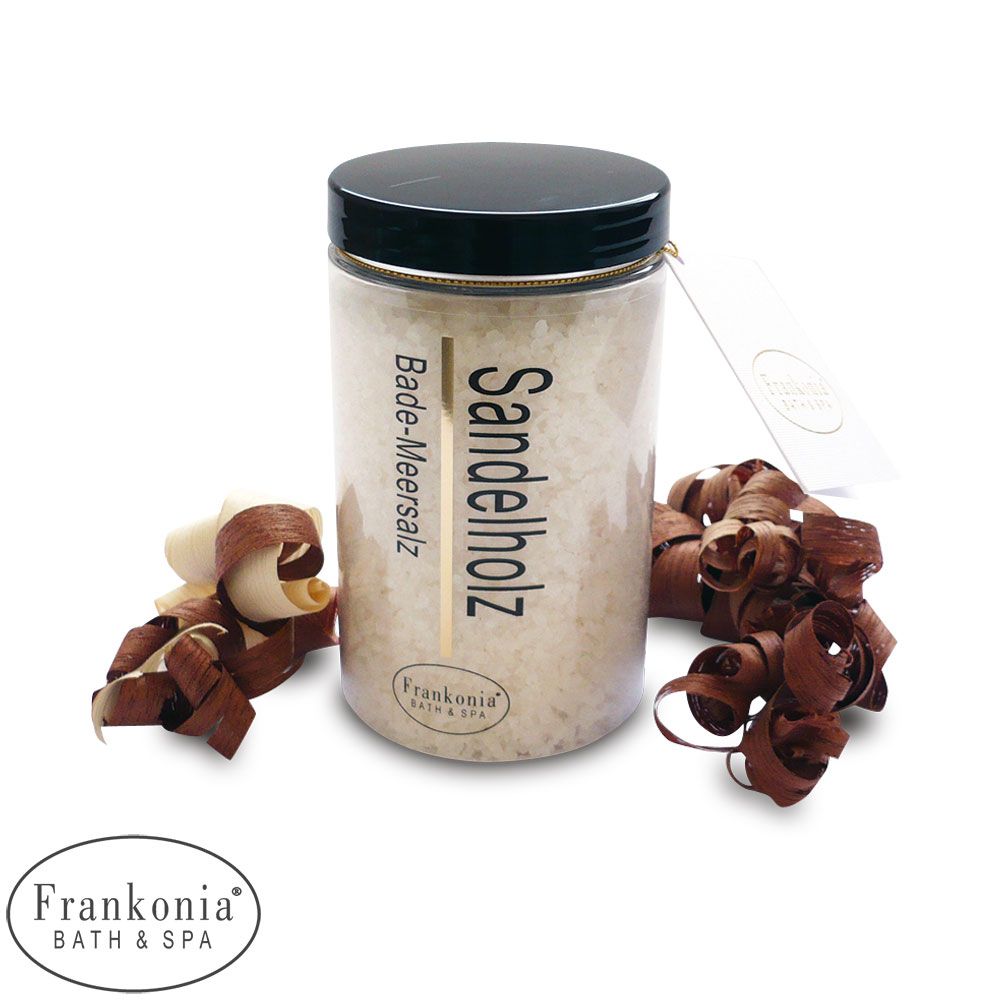 Un pot de sel de bain Frankonia au bois de santal. Le pot transparent révèle le sel et est fermé par un couvercle noir. Des copeaux de bois et une étiquette sont présents. L'étiquette indique "Bade-Meersalz Sandelholz".