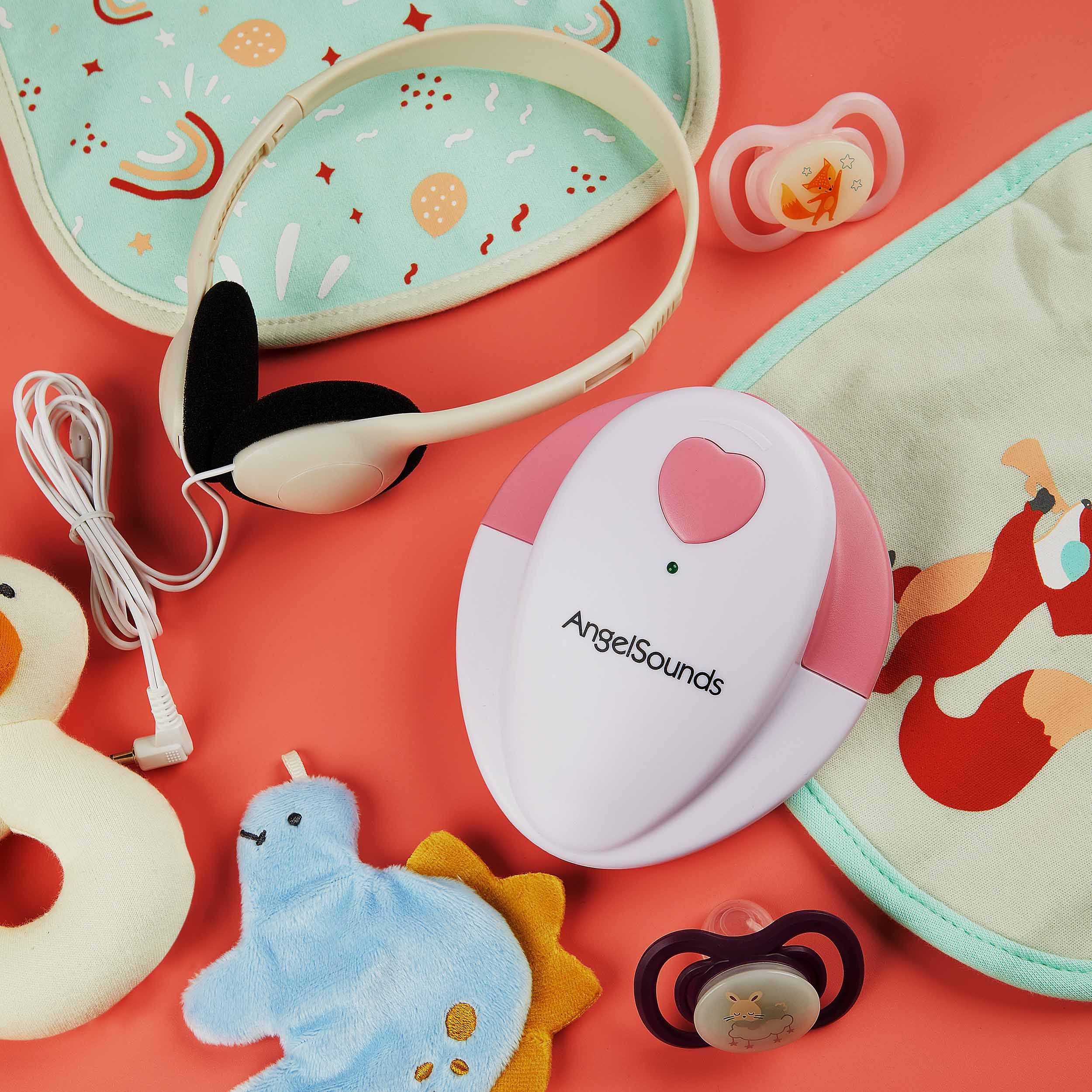Zubehör: Kopfhörer, Schnuller, Lätzchen, Spielzeug und AngelSounds-Gerät. Rosa Hintergrund.