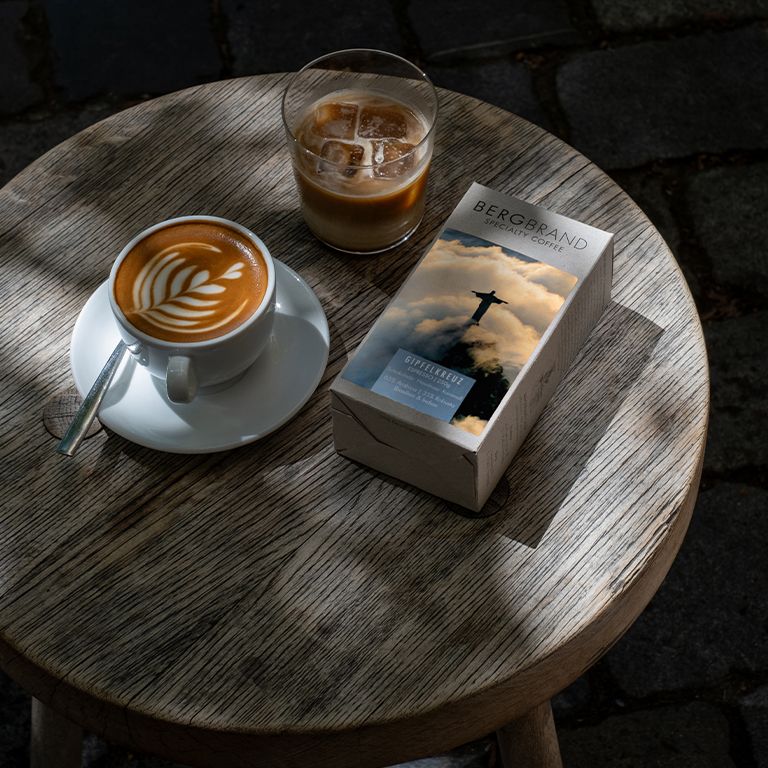 Kaffee und Eisgetränk auf einem Tisch. Daneben eine Packung BERGBRAND Gipfelkreuz Specialty Coffee.