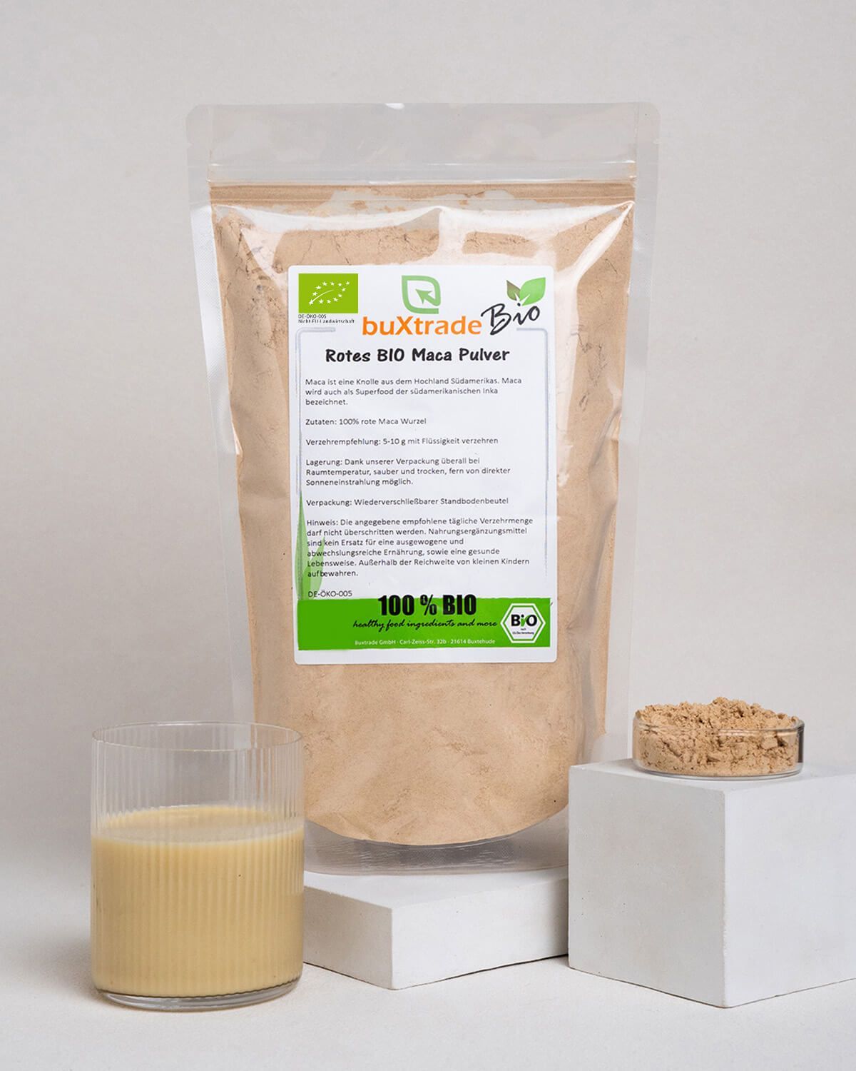 Rotes BIO Maca Pulver in einem Standbodenbeutel, daneben eine kleine Schale mit Pulver und ein Glas mit Getränk.
