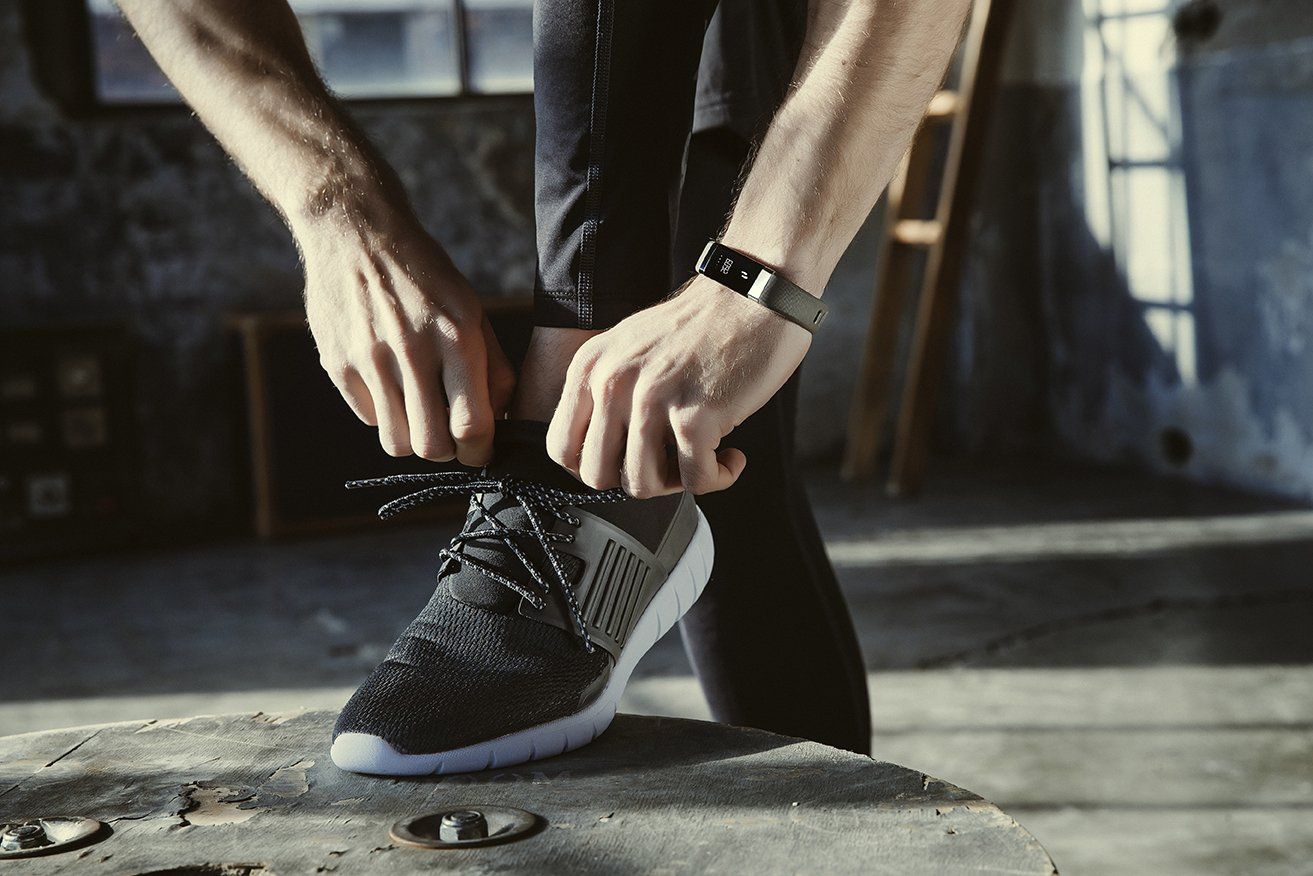 Person bindet Schnürsenkel, trägt schwarzen Fitnesstracker. Grauer Hintergrund, Holzoberfläche.