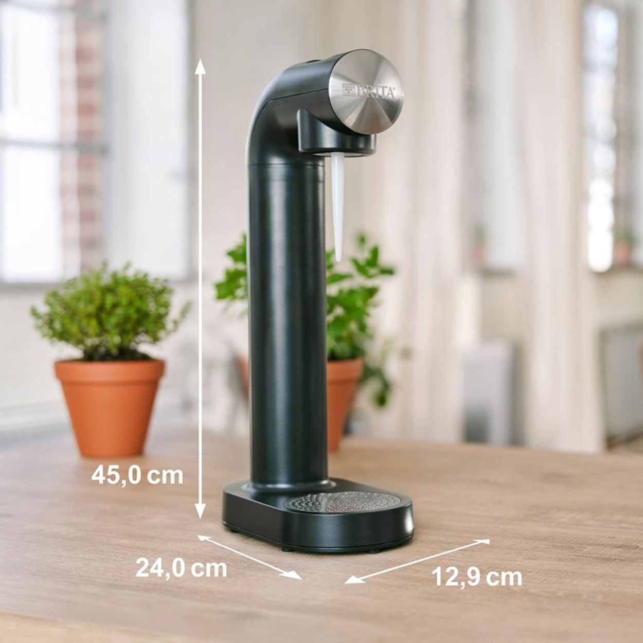 Der schwarze BRITA sodaONE Wassersprudler steht auf einem Holztisch. Die Maße sind 45,0 cm hoch, 24,0 cm breit und 12,9 cm tief. Im Hintergrund sind Pflanzen in Töpfen zu sehen.