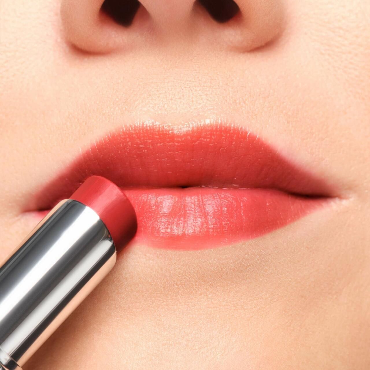 Nahaufnahme von Lippen, die mit einem Lippenbalsam in Korallenrot geschminkt werden. Silberfarbene Hülse.