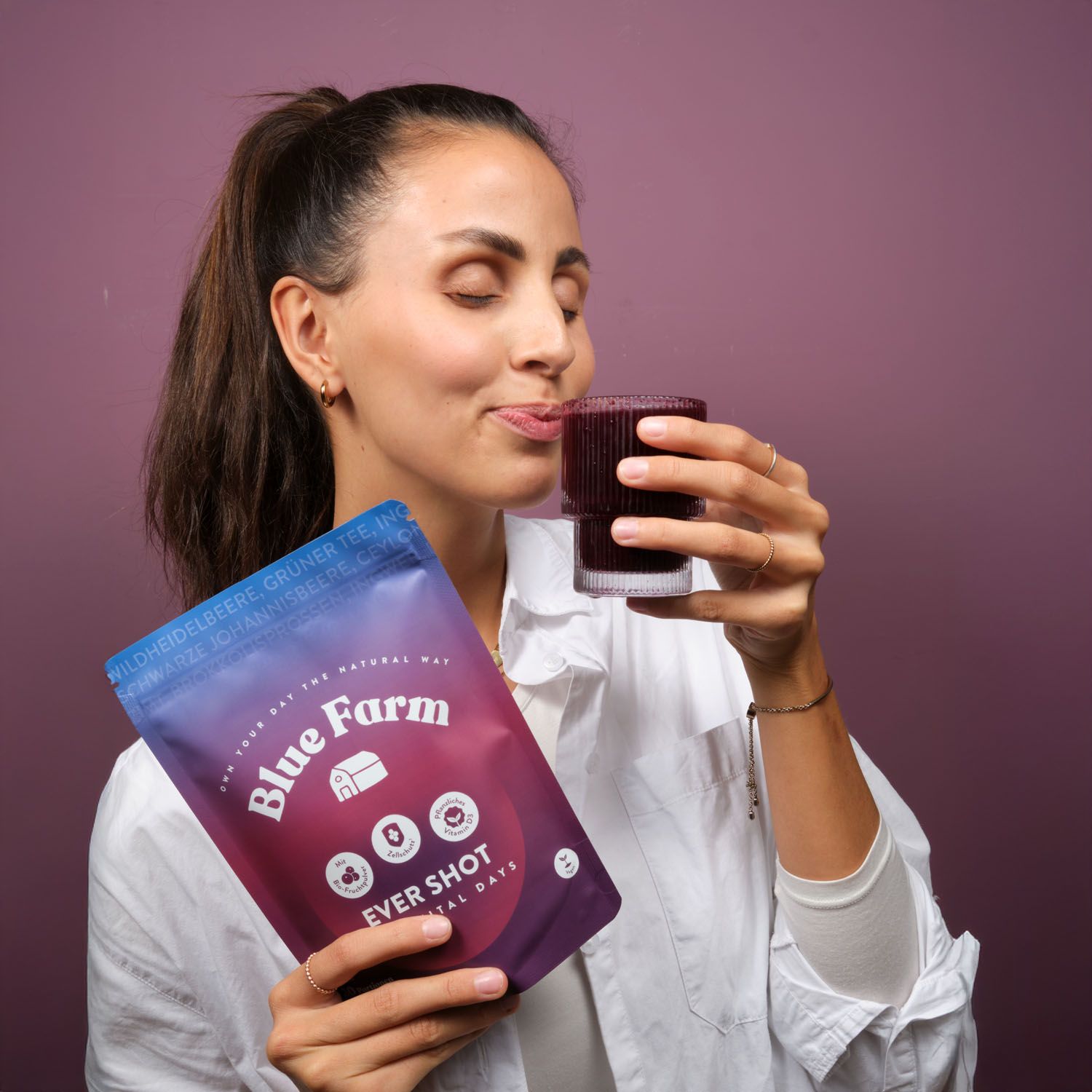 Frau trinkt aus Glas. Hält Beutel "Blue Farm Ever Shot". Logo und Text "More Vital Days" sichtbar. Weißes Hemd.