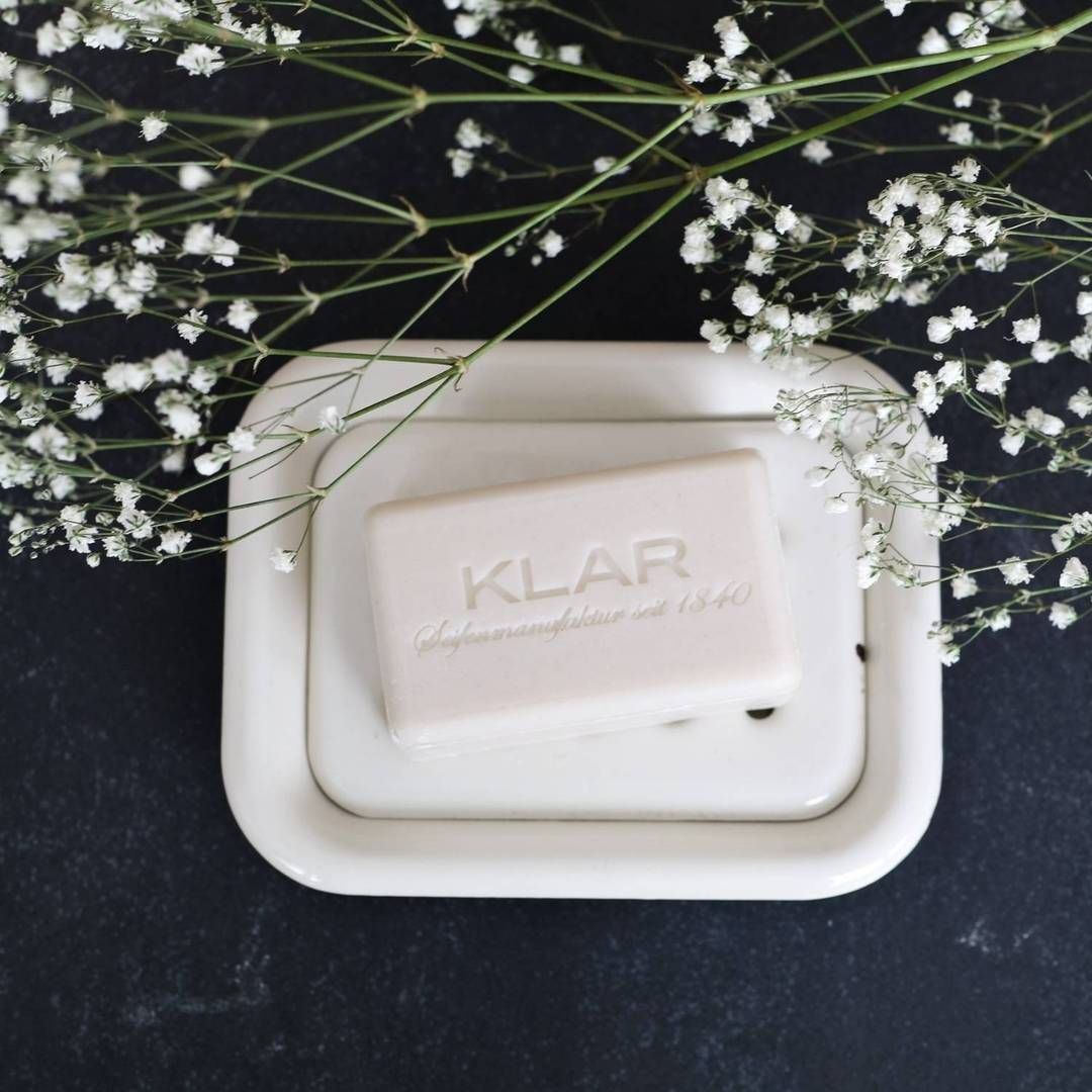 Savon crème avec logo Klar sur porte-savon émaillé blanc. Décoration de fleurs blanches.