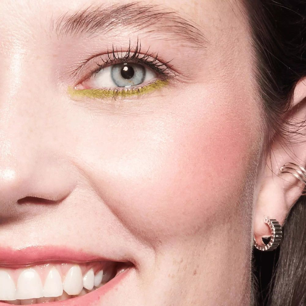 Nahaufnahme eines Gesichts mit Rouge auf den Wangen. Sie trägt Ohrringe und Eyeliner. Helles Make-up.