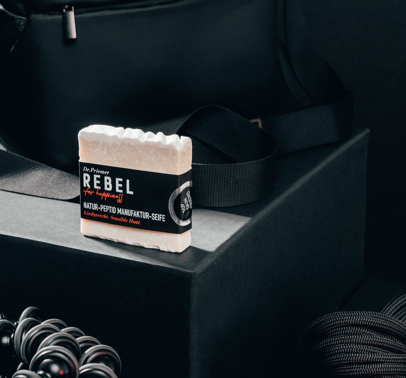 Seife mit schwarzem Etikett. Aufschrift: Dr. Priemer, REBEL for happiness, Natur-Peptid Manufaktur-Seife, kindgerecht, sensible Haut. Auf schwarzem Podest.