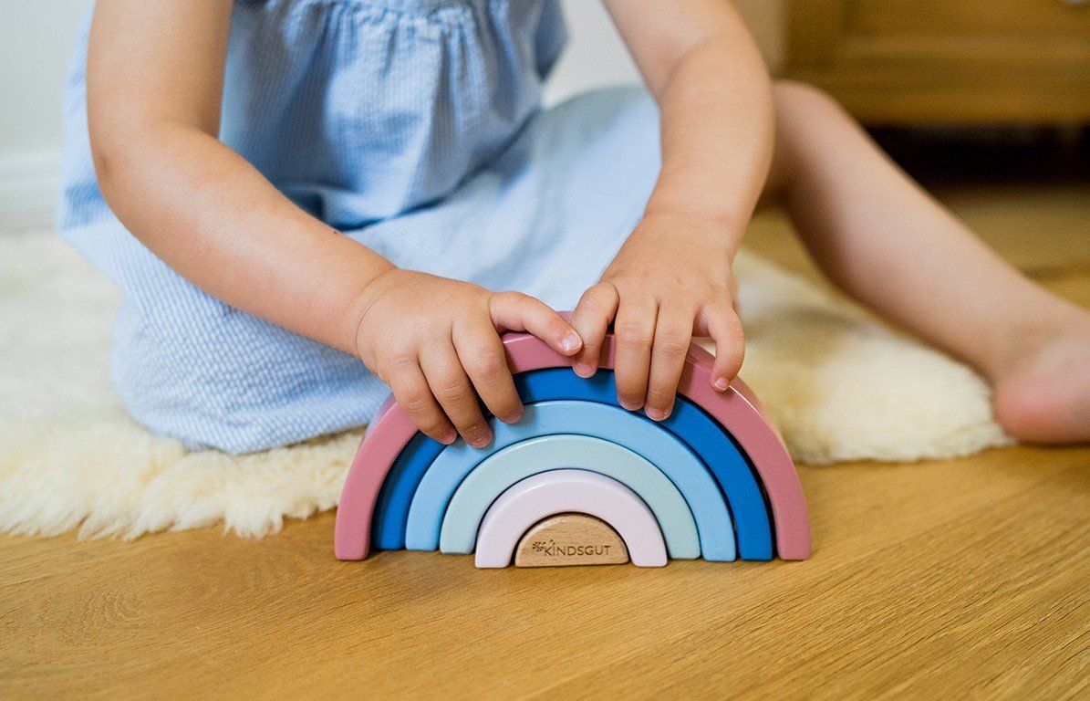 Kind spielt mit einem Holz-Regenbogen. Das Spielzeug besteht aus sieben farbigen Bögen. Das Kind sitzt auf einem Schaffell.