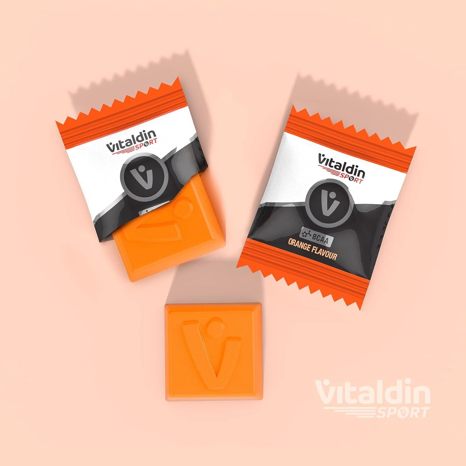 Drei orangefarbene Gummibärchen. Zwei in geöffneten Verpackungen, eines lose. Marke VITALDIN SPORT.