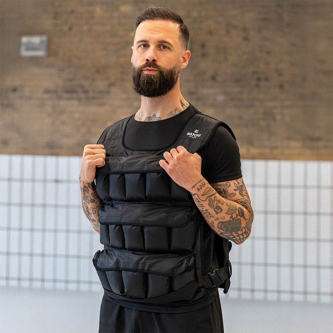 Mann trägt schwarze Gewichtsweste. Marke Matchu Sports. Hintergrund: Fitnessstudio.