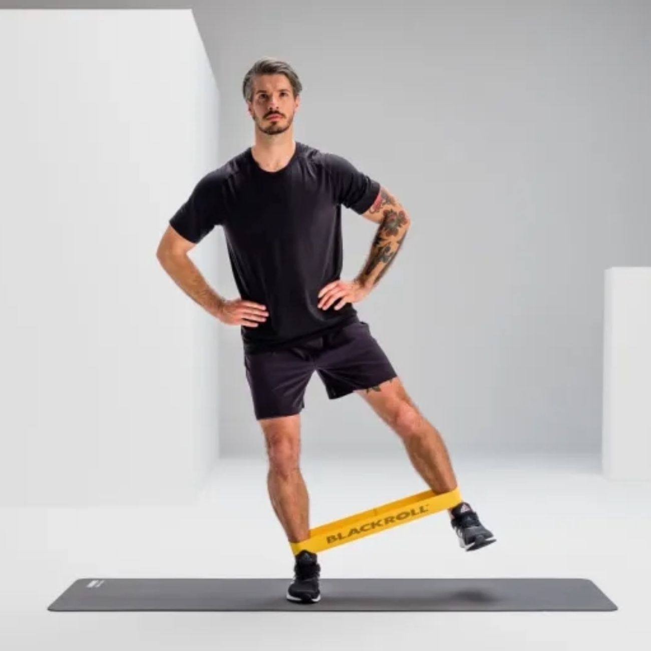 Mann trainiert mit gelbem Loop Band. Band um das Bein. Aufschrift BLACKROLL. Auf grauer Matte. Schwarze Kleidung.