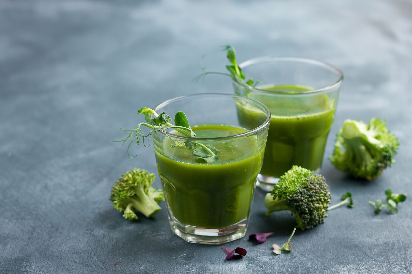 Zwei Gläser grüner Flüssigkeit mit Sprossen, Brokkoli-Röschen auf grauem Untergrund.