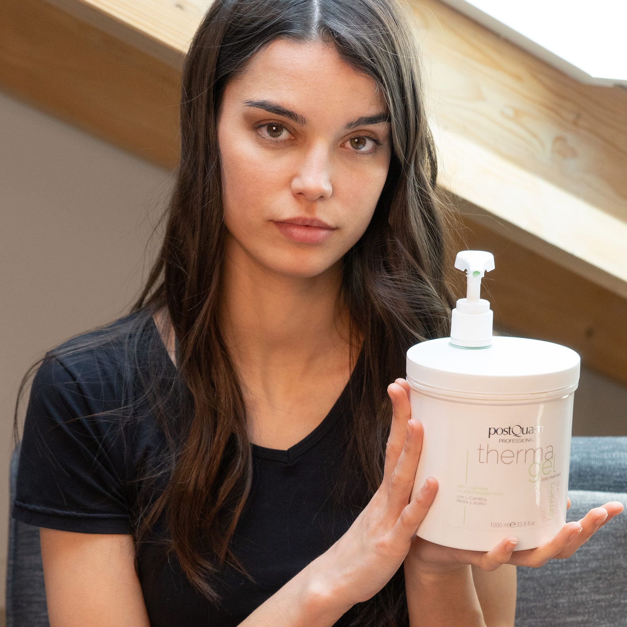 Frau hält weißen Behälter 'PostQuam therma gel' mit Pumpe. Frau trägt schwarzes Oberteil.