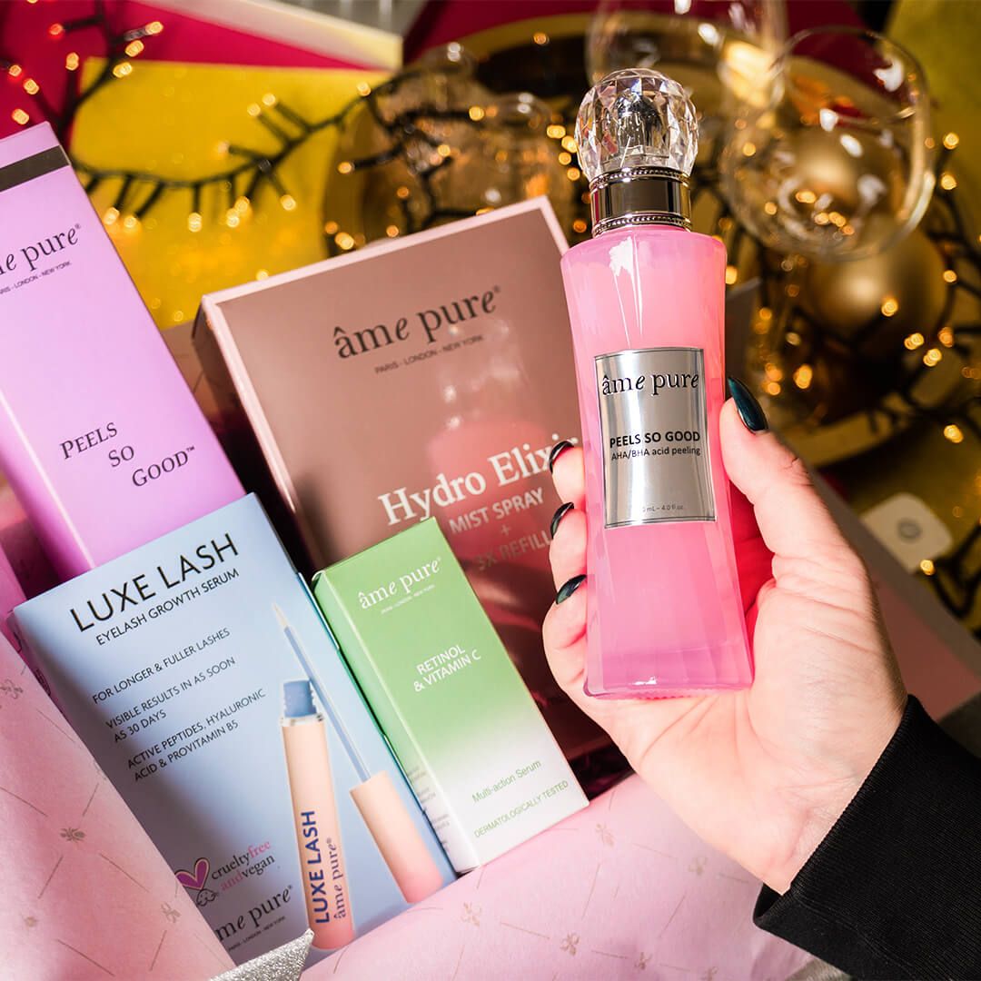 Een hand houdt een roze fles met zilveren etiket vast. Andere producten zijn te zien in een geschenkverpakking, waaronder een wimperserum.