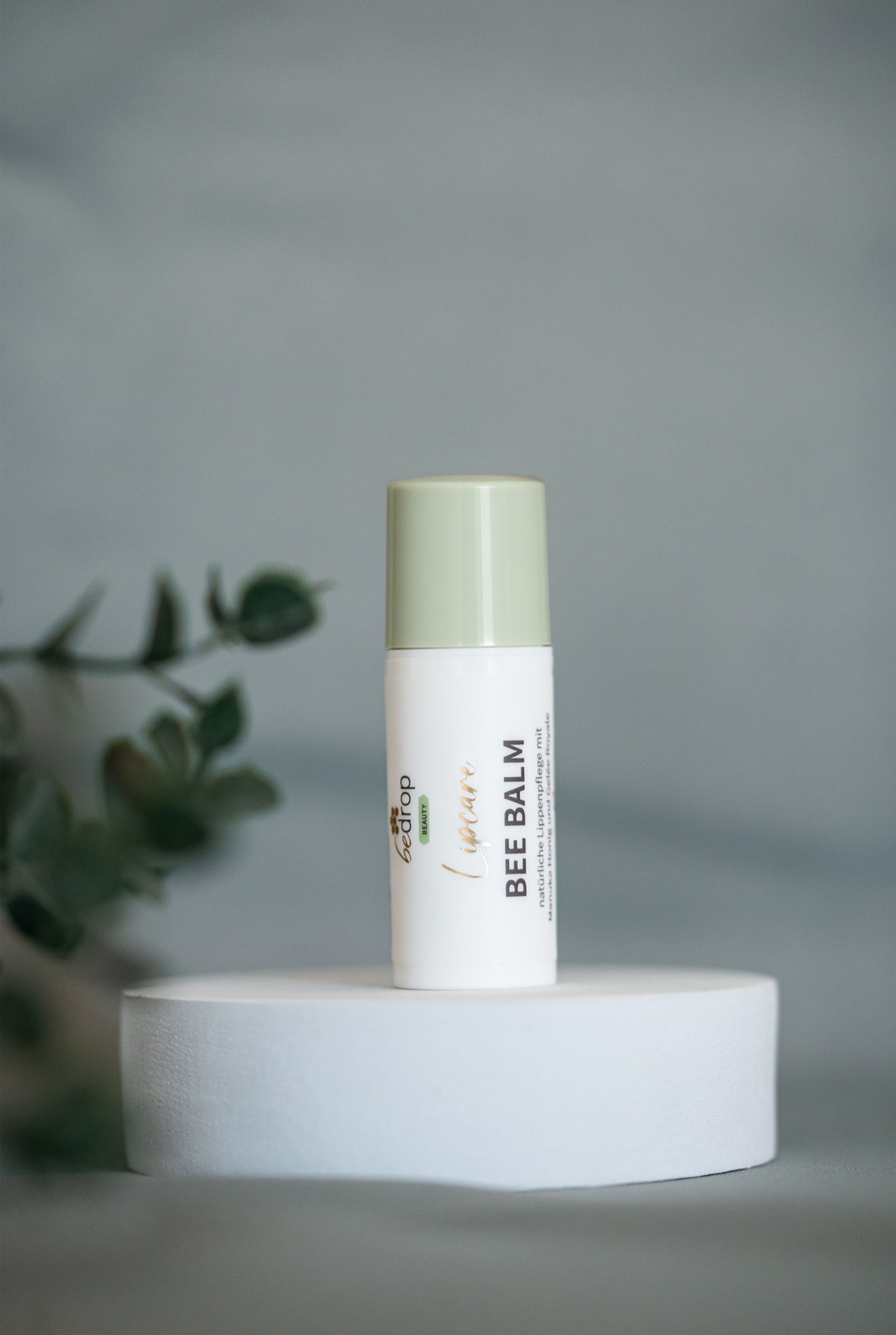Weißer Lippenbalsam mit grünem Deckel auf weißem Podest. Aufschrift: BEE BALM, Lipcare, bedrop.