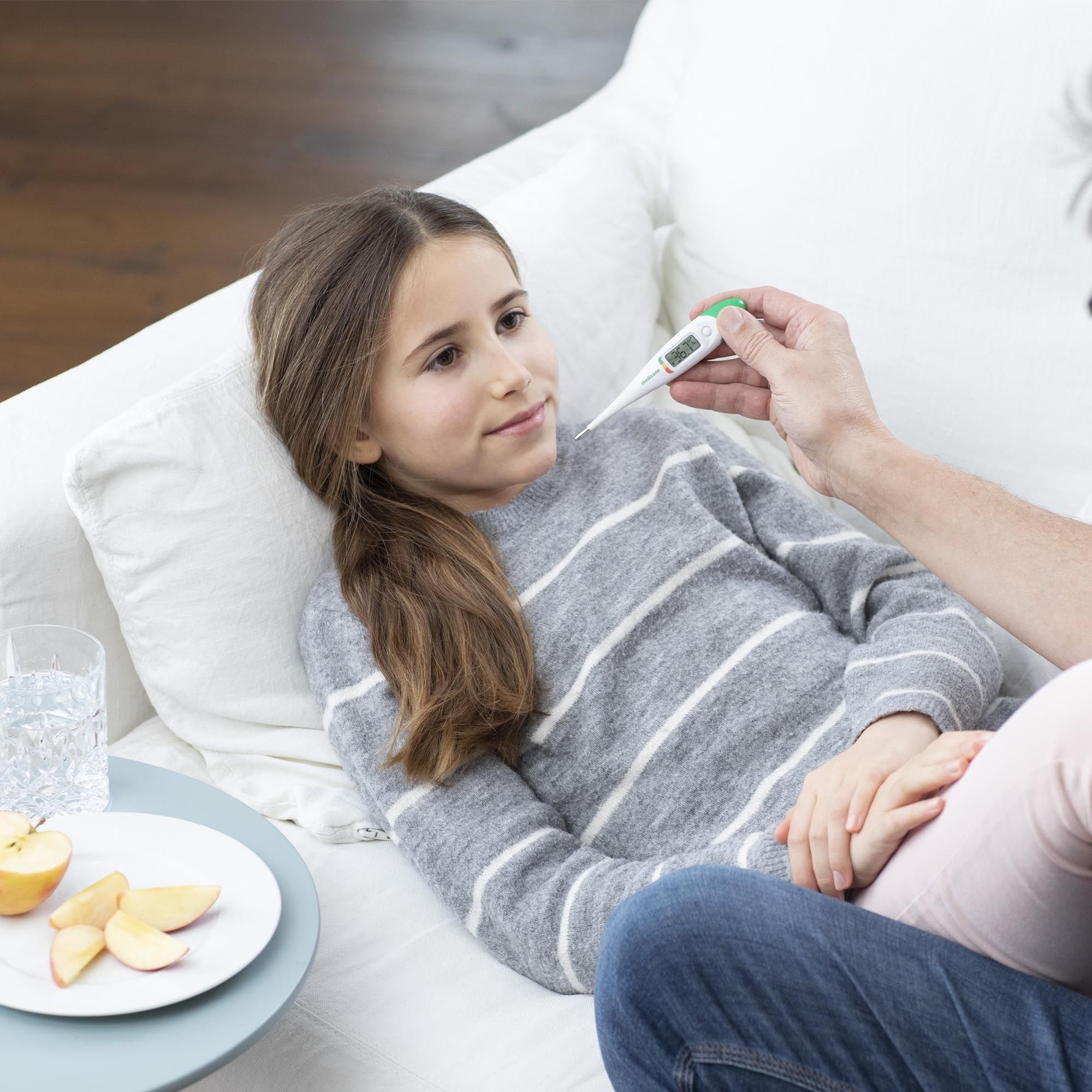 Kind liegt auf Sofa, Fieberthermometer im Mund. Person hält Thermometer. Neben dem Kind: Wasser, Apfel.
