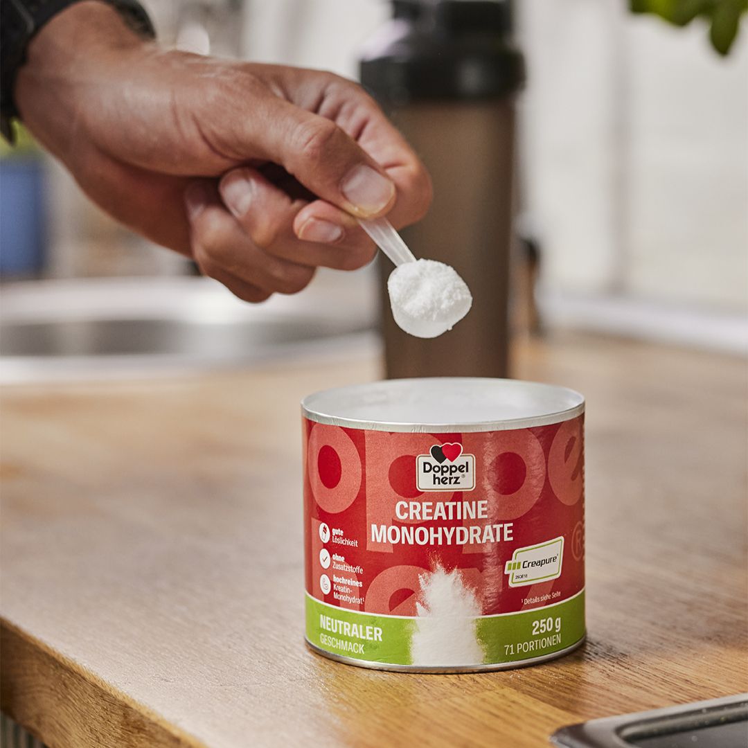 Dose mit rotem Etikett, Produktname und Creapure-Logo. Löffel mit weißem Pulver. Hand hält Löffel über Dose.