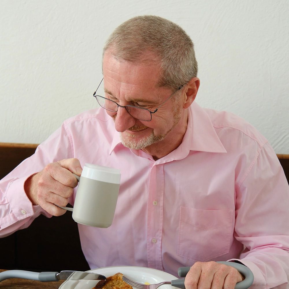 Älterer Mann trinkt aus einem grauen Becher mit großem Henkel. Er sitzt am Tisch, isst.