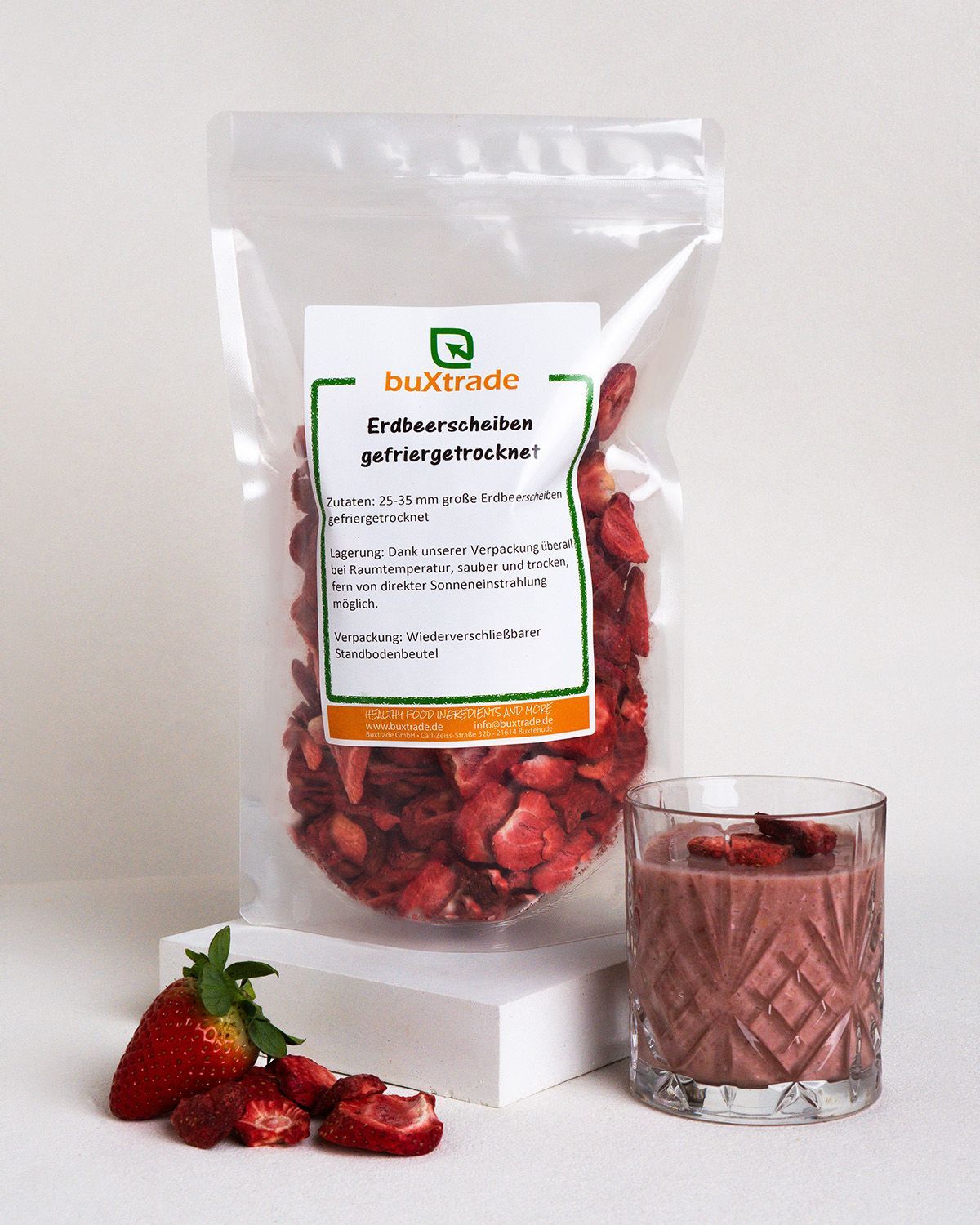 Transparente Beutel mit gefriergetrockneten Erdbeerscheiben. Glas mit Erdbeersmoothie. Frische Erdbeeren und Scheiben daneben.