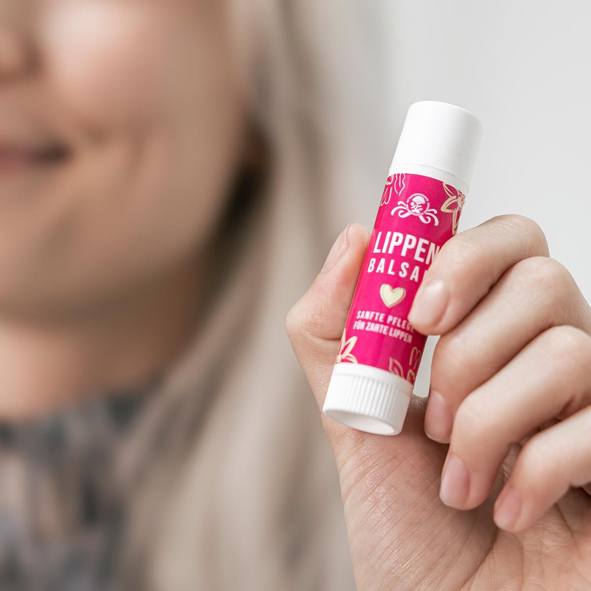 Person hält einen Lippenbalsam-Stift. Pinkfarbene Hülle mit Aufschrift: LIPPEN BALSAM. Weißer Deckel. Hintergrund: verschwommene Person.
