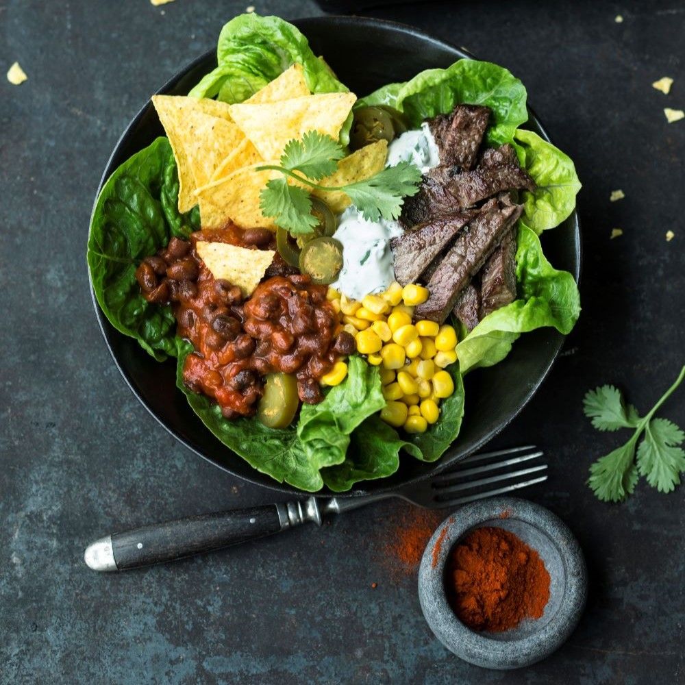 Schwarze Schüssel mit Salat, Fleisch, Mais, Tortilla-Chips, Bohnen, Gurken und Kräutern. Roter Gewürzpulver in Schale.
