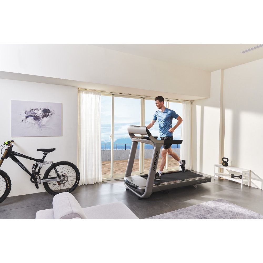 Graues Laufband in Wohnraum. Läufer trainiert. Sichtbar: Fenster, Fahrrad, Hantel, Sofa. Horizon-Logo am Laufband.
