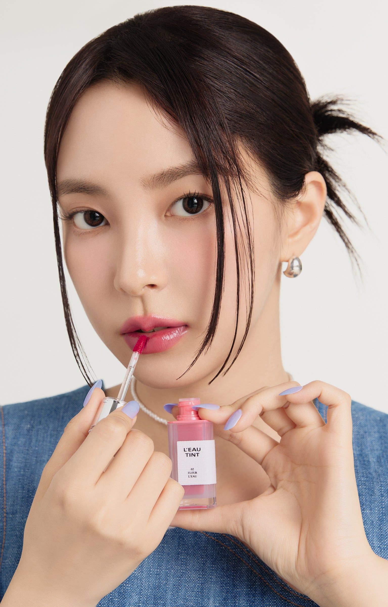 Frau hält BBIA L'eau Tint 02 Fleur L'eau. Sie trägt Jeans-Oberteil und trägt Lippenstift auf.