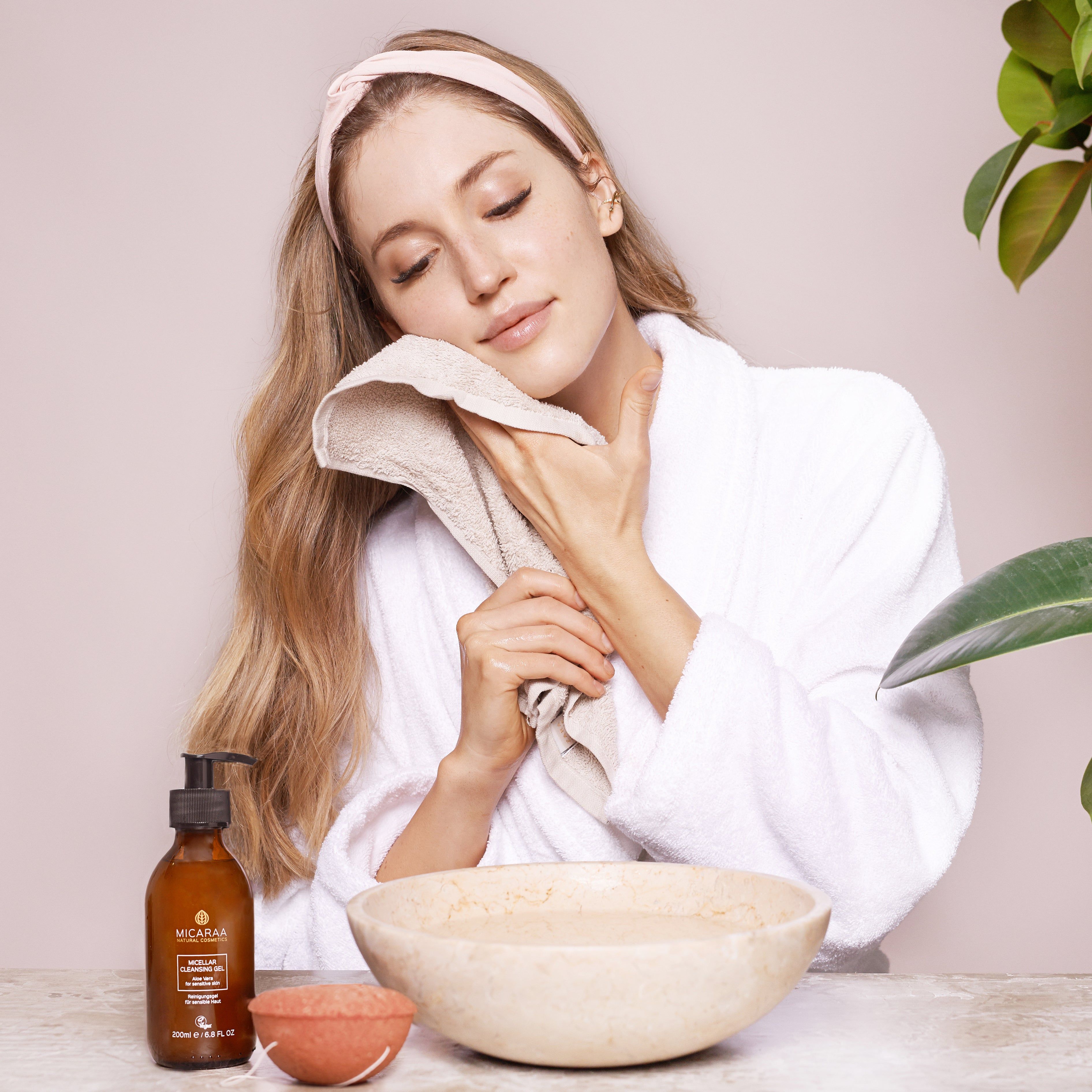 Frau mit Handtuch im Bademantel. Auf dem Tisch: Flasche mit Pumpverschluss, Schale mit Wasser, Schale mit rotem Inhalt. Produkt: MICARAA.