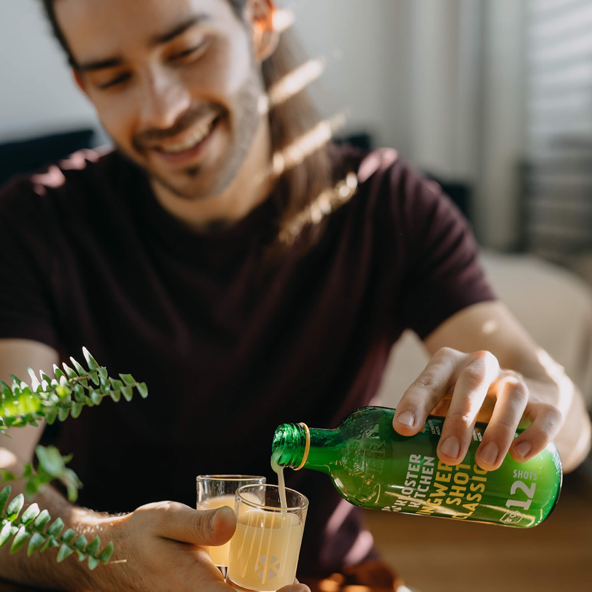 Mann gießt gelbes Getränk aus einer grünen Flasche in zwei Gläser. Flasche: Kloster Kitchen Classic, 12 Shots.