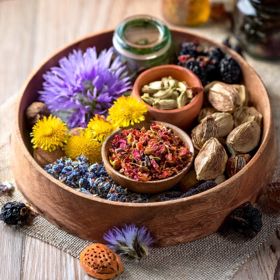 Holzschale mit verschiedenen Kräutern, Blüten und Samen. Mehrere kleine Schalen und Gläser enthalten die Zutaten.
